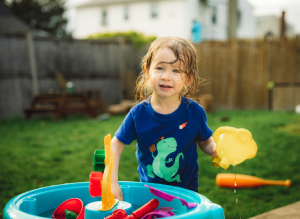 Child playing with toys in a plastic bin filled with water in the foreground, with green grass yard in the background