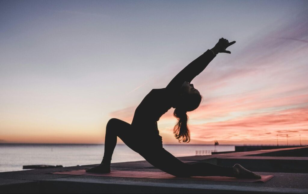 If the Body Doesn’t Keep the Score, What Does? 2 Person silhouetted in a low lunge yoga pose, on the waterfront with a soft pastel sunset in the background.