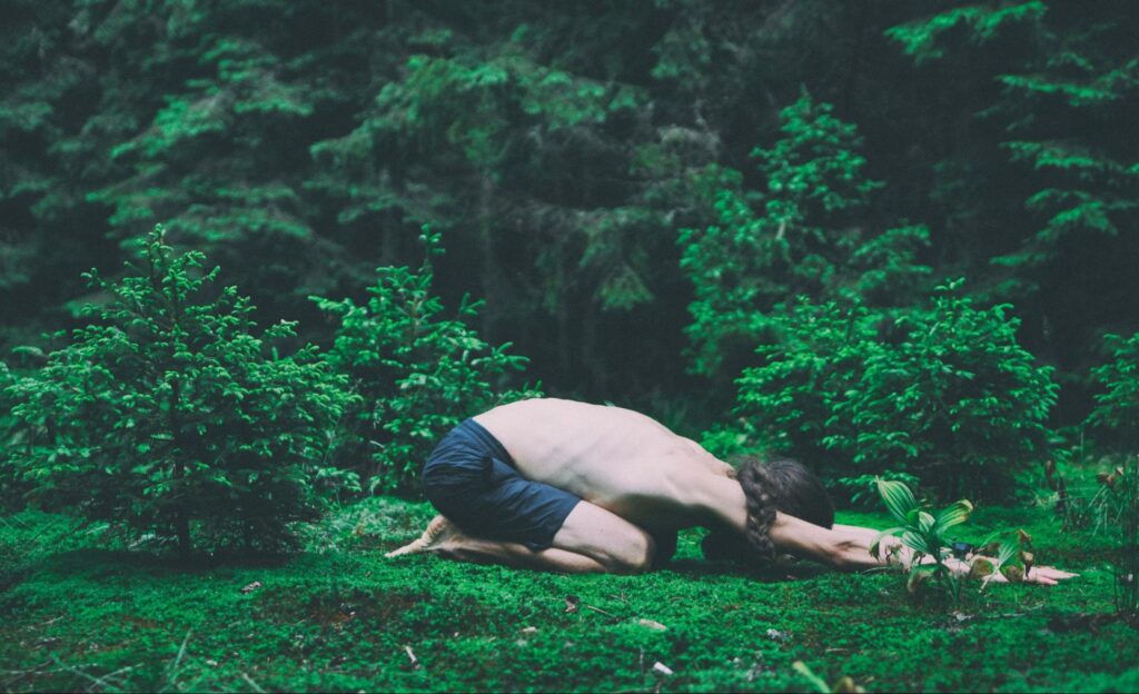 If the Body Doesn’t Keep the Score, What Does? 1 Person in yoga child’s pose with arms outstretched in foreground, with a vibrant green forest surrounding them.